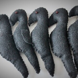 May include: Five black felt crow figurines with red bead eyes. The crows are arranged in a row, facing the right side of the image.