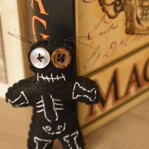 May include: Black felt voodoo doll with white stitching and button eyes. The doll is leaning against a book titled "Magic".