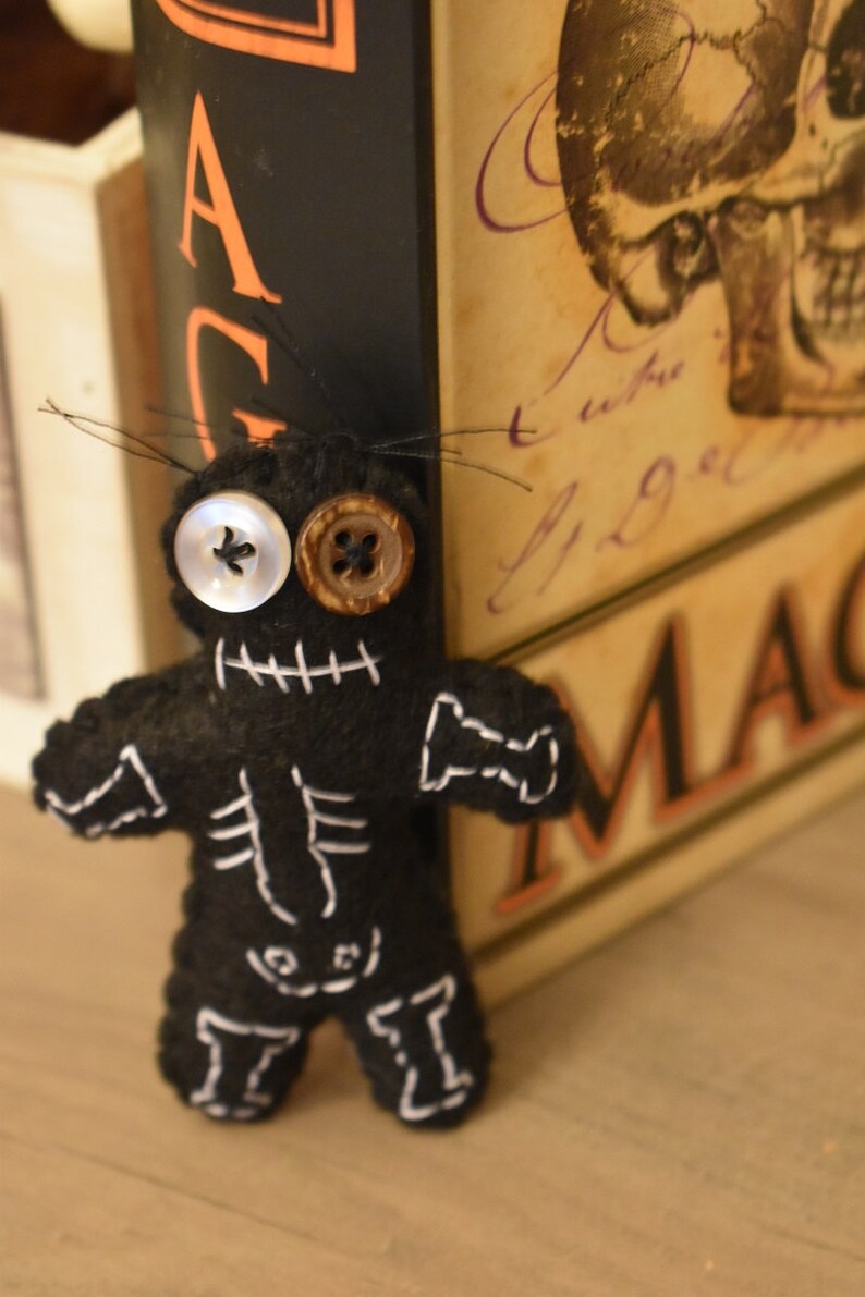 May include: Black felt voodoo doll with white stitching and button eyes. The doll is leaning against a book titled "Magic".