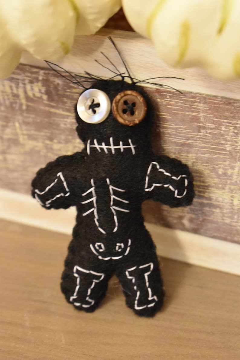 May include: Black felt voodoo doll with white stitching, button eyes, and a stitched-on mouth. The doll has a skeleton design stitched on the front.