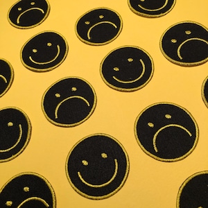May include: A yellow background with black and yellow smiley face and frowny face patches. The patches are circular and have a yellow outline.