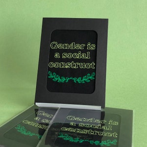 May include: A black card with a white frame and green leaves around the text "Gender is a social construct" in yellow.
