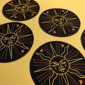 Peut inclure: Cinq patchs thermocollants brodés noirs avec un soleil doré au visage pleurant et des cœurs orange. Le soleil a un contour ondulé et des rayons.