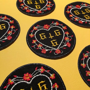 Puede incluir: Parches bordados negros circulares con un diseño de corazón. El corazón contiene letras doradas y está rodeado por un borde blanco y flores rojas y amarillas. Los parches están dispuestos sobre una superficie amarilla.