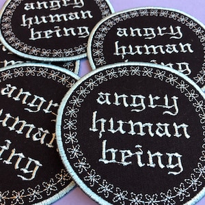 May include: Several black circular patches embroidered with the words "angry human being" in white gothic script. Each patch features a floral border and a light blue edge.