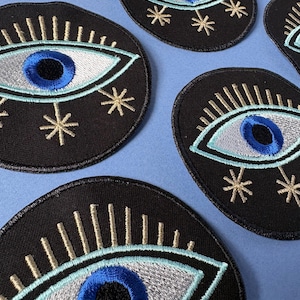Peut inclure: Plusieurs écussons brodés noirs en forme d'œil. L'œil a un iris bleu, des paupières argentées et des cils dorés, entouré d'éclats d'étoiles dorées. Ces écussons sont probablement destinés aux vêtements ou aux accessoires.