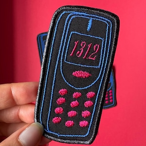 May include: Black embroidered patch in the shape of a retro cell phone with a blue outline and accents. The phone displays the number "1312" in pink. Additional patches are visible in the background against a red backdrop.