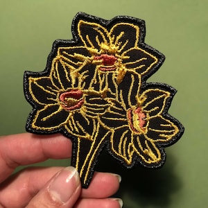 Könnte beinhalten: Ein schwarzer gestickter Aufnäher mit drei gelben Narzissen mit roten Zentren.