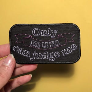 Peut inclure: Patch brodé noir avec des coutures blanches qui disent "Only you can judge me" avec un ruban rose au-dessus du texte.