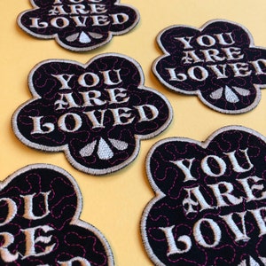 Könnte beinhalten: Fünf schwarze Filz-Blumenpatches mit rosa Nähten an den Rändern. Jeder Patch ist mit weißer Stickerei versehen, auf der "You Are Loved" steht.