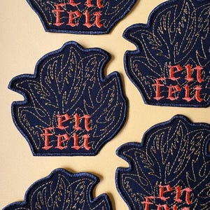 Peut inclure: Plusieurs écussons brodés bleu marine en forme de flamme avec des coutures dorées. Chaque écusson porte l'inscription "en feu" en lettres gothiques rouges. Les écussons sont disposés sur une surface beige clair.