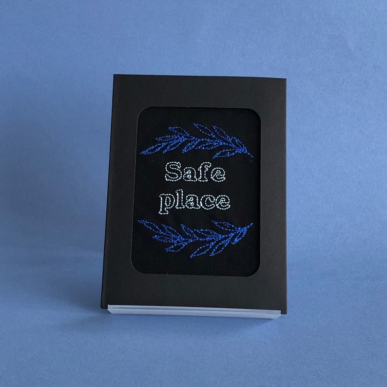 May include: A black box with a white border and a black fabric insert. The fabric has a blue embroidered design of two branches with the words "Safe Place" in white.