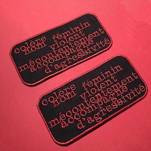 May include: Two black fabric patches with red embroidered text. The text reads "colere feminin nom violent mecontement accompagne d'aggressivite".