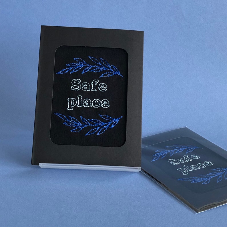 May include: A black card with a white embroidered design of a branch with leaves and the words "Safe Place". The card is displayed on a blue surface.