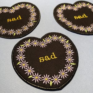 Peut inclure: Trois patchs en forme de cœur en feutre noir avec une bordure de chaîne de marguerites violettes. Chaque patch a le mot "sad" brodé en fil jaune.