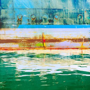 May include: A man in red swim trunks stands on a wooden platform in front of a blue and green body of water. The platform has a ladder leading into the water. The background is a weathered blue and green wall with a row of round portholes.