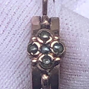 K&ouml;nnte beinhalten: Nahaufnahme eines goldfarbenen Ohrrings mit floralem Design. Der Ohrring hat eine kleine, verzierte Blume mit f&uuml;nf dunkelgr&uuml;nen Steinen in der Mitte. Der Ohrring hat einen Hakenverschluss.