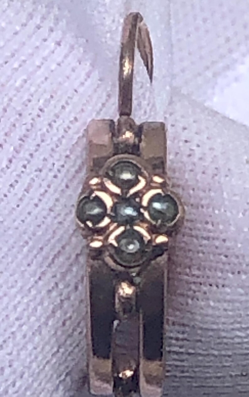 K&ouml;nnte beinhalten: Nahaufnahme eines goldfarbenen Ohrrings mit floralem Design. Der Ohrring hat eine kleine, verzierte Blume mit f&uuml;nf dunkelgr&uuml;nen Steinen in der Mitte. Der Ohrring hat einen Hakenverschluss.