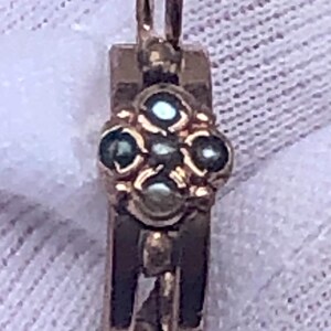 K&ouml;nnte beinhalten: Ein goldfarbener Anh&auml;nger mit floralem Design. Der Anh&auml;nger hat oben eine Schlaufe zum Aufh&auml;ngen und einen rechteckigen K&ouml;rper. Das florale Design besteht aus vier dunkel gef&auml;rbten Steinen. Der Anh&auml;nger ist wahrscheinlich aus Gold.