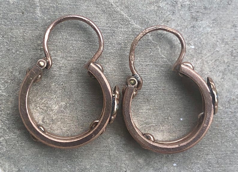 K&ouml;nnte beinhalten: Ein Paar antike Creolen aus Ros&eacute;gold. Jeder Ohrring hat einen Klappverschluss und kleine dekorative Elemente. Die Ohrringe werden vor einem strukturierten, grauen Hintergrund pr&auml;sentiert, der ihren Vintage-Charme und ihr Design hervorhebt.