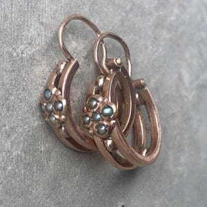 K&ouml;nnte beinhalten: Ein Paar antike Creolen aus Ros&eacute;gold. Jede Ohrring hat ein Blumenmuster mit kleinen, runden, grau-blauen Edelsteinen. Die Ohrringe haben einen klassischen B&uuml;gelverschluss und werden auf einer grauen Oberfl&auml;che pr&auml;sentiert.