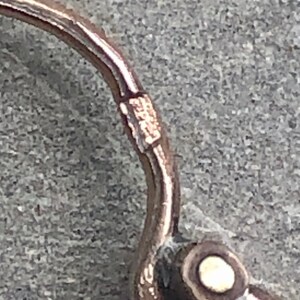 K&ouml;nnte beinhalten: Nahaufnahme eines ros&eacute;goldenen Ohrringverschlusses. Der Ohrring hat ein geschwungenes Design mit einem kleinen, runden, wei&szlig;en Akzent. Der Verschluss ist mit kleinen Markierungen versehen.