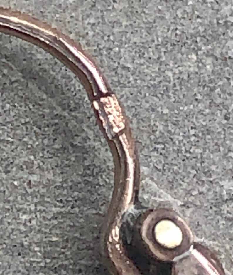 K&ouml;nnte beinhalten: Nahaufnahme eines ros&eacute;goldenen Ohrringverschlusses. Der Ohrring hat ein geschwungenes Design mit einem kleinen, runden, wei&szlig;en Akzent. Der Verschluss ist mit kleinen Markierungen versehen.