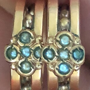 K&ouml;nnte beinhalten: Nahaufnahme von zwei goldenen Ringen mit floralem Design. Jeder Ring hat eine Ansammlung runder, hellblauer Edelsteine in einer Goldfassung. Die Ringe haben ein poliertes, gl&auml;nzendes Aussehen.