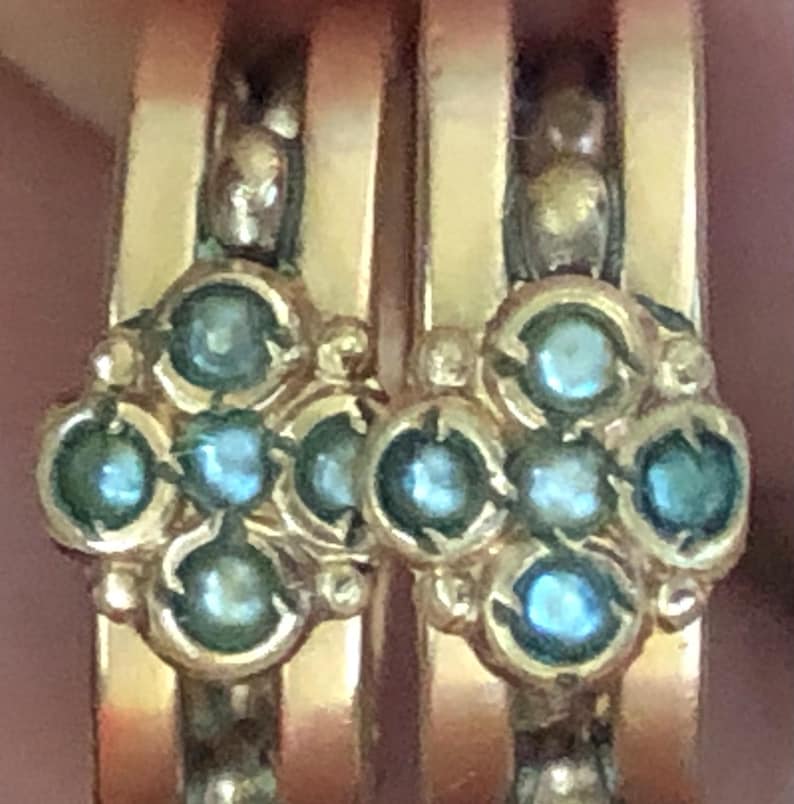 K&ouml;nnte beinhalten: Nahaufnahme von zwei goldenen Ringen mit floralem Design. Jeder Ring hat eine Ansammlung runder, hellblauer Edelsteine in einer Goldfassung. Die Ringe haben ein poliertes, gl&auml;nzendes Aussehen.