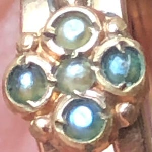 K&ouml;nnte beinhalten: Nahaufnahme eines goldfarbenen Rings mit einem Cluster aus f&uuml;nf kleinen, runden, hellgr&uuml;nen Edelsteinen. Die Edelsteine sind in einem floralen oder sternf&ouml;rmigen Muster angeordnet und haben eine helle, reflektierende Qualit&auml;t. Der Ring ist sichtbar.