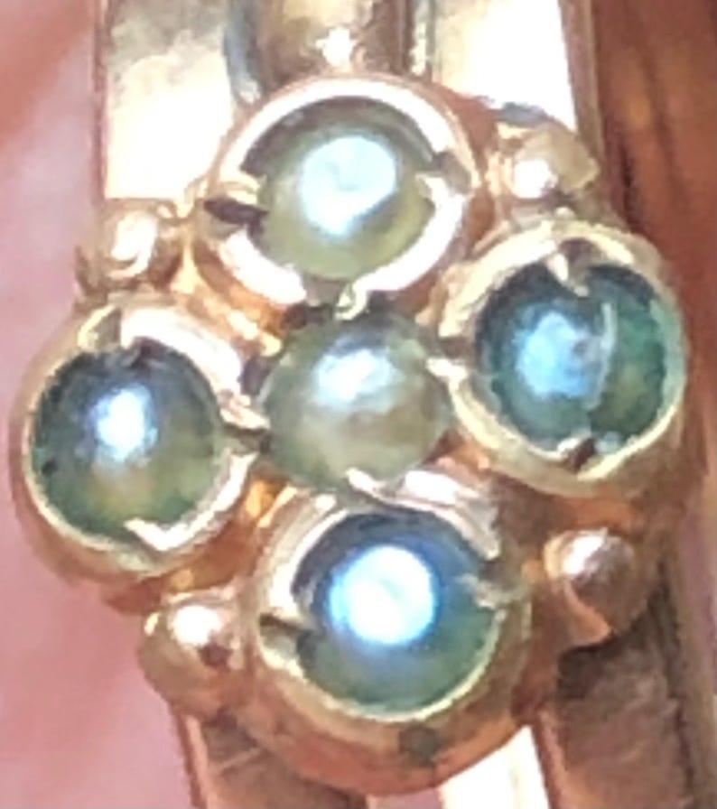 K&ouml;nnte beinhalten: Nahaufnahme eines goldfarbenen Rings mit einem Cluster aus f&uuml;nf kleinen, runden, hellgr&uuml;nen Edelsteinen. Die Edelsteine sind in einem floralen oder sternf&ouml;rmigen Muster angeordnet und haben eine helle, reflektierende Qualit&auml;t. Der Ring ist sichtbar.