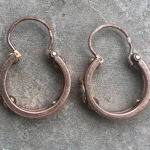 K&ouml;nnte beinhalten: Ein Paar antike Creolen. Die Ohrringe sind aus einem kupferfarbenen Metall gefertigt und weisen kleine dekorative Elemente auf. Die Reifen haben einen Klappverschluss. Die Ohrringe werden auf einer grauen Oberfl&auml;che pr&auml;sentiert.