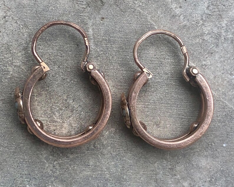 K&ouml;nnte beinhalten: Ein Paar antike Creolen. Die Ohrringe sind aus einem kupferfarbenen Metall gefertigt und weisen kleine dekorative Elemente auf. Die Reifen haben einen Klappverschluss. Die Ohrringe werden auf einer grauen Oberfl&auml;che pr&auml;sentiert.