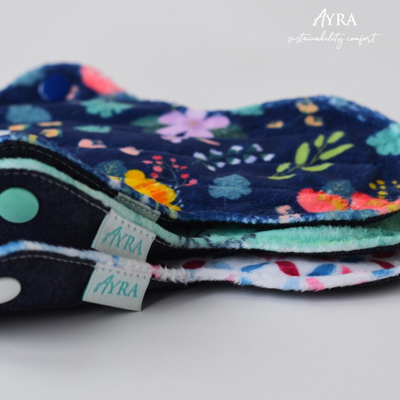 Puede incluir: Primer plano de varias compresas menstruales de tela reutilizables. Las compresas presentan un estampado floral azul marino, un borde verde menta y una etiqueta blanca con el nombre de la marca "Ayra".