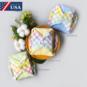 Puede incluir: Tres compresas menstruales de tela reutilizables con estampados de cuadros: azul, amarillo y verde. Cada compresa tiene un cierre de botón a presión y se muestra con ramitas de algodón y el texto "Gingham Blue", "Gingham Yellow" y "Gingham Green".