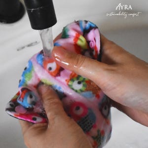 Puede incluir: Un pa&ntilde;o rosa y negro con estampado de b&uacute;hos se lava con agua corriente. El pa&ntilde;o presenta ilustraciones coloridas de b&uacute;hos en varios colores. El texto "AYRA sustainability comfort" es visible en la esquina superior derecha.