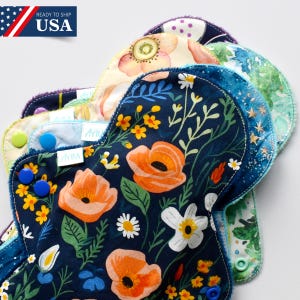 Puede incluir: Una pila de compresas menstruales de tela reutilizables con coloridos estampados florales. Las compresas presentan una variedad de diseños, incluyendo amapolas naranjas, margaritas y otros elementos botánicos. Las compresas tienen cierres de presión. El texto 'READY TO SHIP USA' es visible.