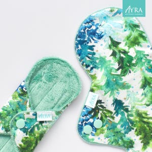Peut inclure: Deux serviettes hygiéniques lavables en tissu avec un imprimé forêt à l'aquarelle dans les tons de vert et de bleu. Les serviettes ont un intérieur vert menthe doux et des fermetures à pression. La marque "Ayra" est visible.