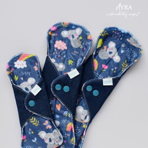 Puede incluir: Tres compresas menstruales de tela reutilizables con exterior de mezclilla azul oscuro e interior estampado con koalas, arco&iacute;ris y elementos florales. Cada compresa tiene un cierre de bot&oacute;n a presi&oacute;n y una peque&ntilde;a etiqueta blanca con la marca "AYRA".