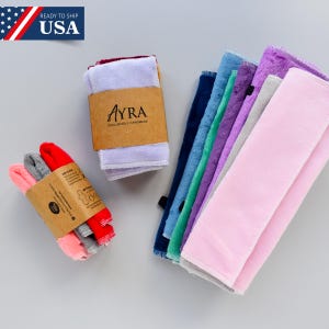 May include: A collection of colorful, soft face cloths in various shades, including pink, purple, blue, and gray. Some are bundled with brown paper bands that feature the brand name "Ayra" and the text "Exclusively Handmade". The image also includes a "Ready to Ship USA" banner.