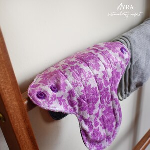 Puede incluir: Una compresa menstrual reutilizable con un estampado floral morado y broches morados. La compresa est&aacute; hecha de tela suave y absorbente, dise&ntilde;ada para la comodidad y la sostenibilidad. Cuelga de un estante de madera.