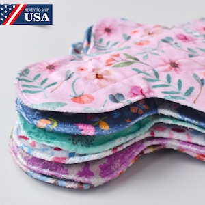 Puede incluir: Una pila de compresas de tela reutilizables. Las compresas presentan estampados florales en rosa, azul, verde y morado. Están hechas de tela suave y absorbente, diseñadas para la comodidad y la sostenibilidad.