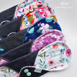 Puede incluir: Una pila de compresas menstruales de tela reutilizables con varios estampados florales y de b&uacute;hos. Las compresas tienen un reverso de mezclilla oscura y se aseguran con broches. La imagen incluye el texto "AYRA sustainability comfort".