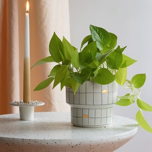 Peut inclure: Un pot en céramique blanche avec un motif en grille et des accents dorés contient une plante verte en pot. Le pot est posé sur une table blanche mouchetée à côté d'un bougeoir blanc avec une bougie allumée.