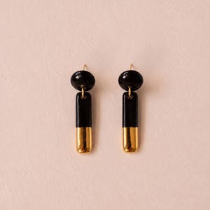 Puede incluir: Un par de pendientes colgantes con una pequeña esfera negra en la parte superior conectada a un colgante rectangular vertical. El colgante es negro en la mitad superior y dorado metálico en la mitad inferior, con un acabado liso y brillante.
