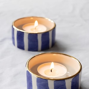 May include: Two white ceramic candle holders with blue and gold stripes. Each holder has a lit tea light candle inside.