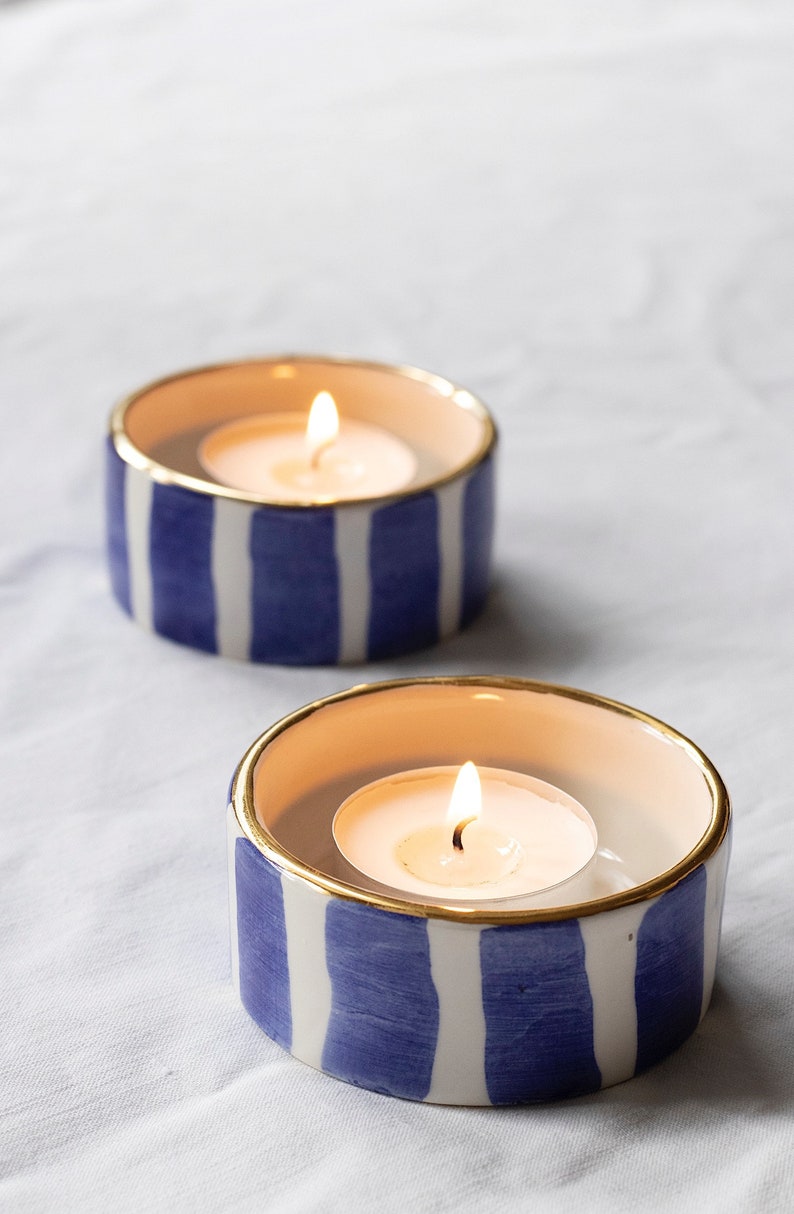 May include: Two white ceramic candle holders with blue and gold stripes. Each holder has a lit tea light candle inside.