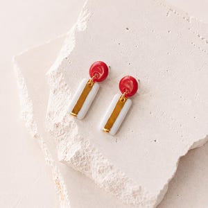 Pode incluir: Um par de brincos pendentes com um pino circular vermelho brilhante ligado a um pendente retangular branco. O pendente branco tem uma faixa metálica vertical de cor dourada no centro. Os brincos são apresentados como um conjunto.