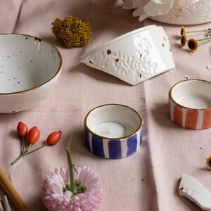 May include: A collection of white ceramic bowls with gold accents, including a small bowl with a blue and white striped pattern, a larger bowl with a red and white striped pattern, and a larger bowl with a white and gold pattern. The bowls are arranged on a pink linen tablecloth with flowers and other decorative elements.