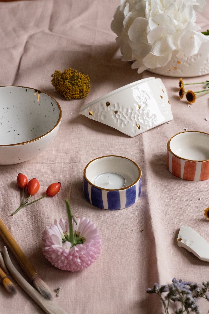 May include: A collection of white ceramic bowls with gold accents, including a small bowl with a blue and white striped pattern, a larger bowl with a red and white striped pattern, and a larger bowl with a white and gold pattern. The bowls are arranged on a pink linen tablecloth with flowers and other decorative elements.